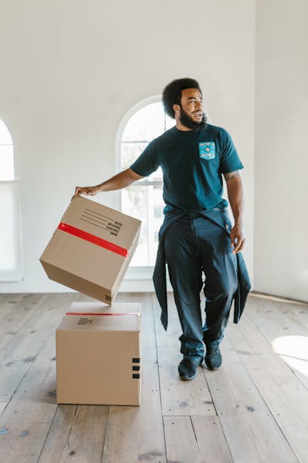 A man with dark curly hair and a beard, dressed in a navy blue T-shirt with a logo on the chest and dark trousers, is inside a bright, empty room with white walls and wooden flooring. He is holding a cardboard box with red and black tape, preparing to place it on top of another closed cardboard box situated on the floor. Behind him, a large arched window allows natural light to illuminate the space, indicating a spacious environment suitable for home relocation or furniture transport. The scene captures the logistics of packing and moving, highlighting the careful handling of packaged belongings by a professional removals service, likely provided by Man with Van Enfield, during a residential transfer or house removal process.
