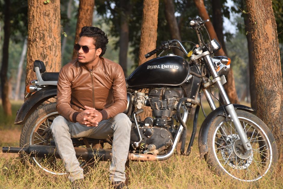 A man with dark hair, wearing sunglasses, a brown leather jacket, and light grey jeans, is sitting on the ground next to a black Royal Enfield motorcycle in a wooded area with tall trees and green foliage. The motorcycle is parked on a patch of grass with its sidestand down, featuring shiny metallic components, a black leather seat, and orange indicator lights. The scene appears to be in natural daylight, with soft shadows cast by the surrounding trees. The man has his hands clasped, looking slightly to the side, and the motorcycle is upright, ready for transport or storage. This setting showcases a casual outdoor environment, possibly related to the preparation or maintenance of a motorbike as part of a house removal or relocation process, as supported by the context of professional removal services offered by Man with Van Enfield.