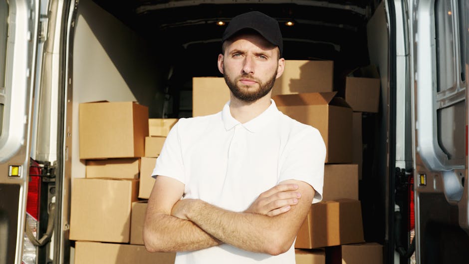 A man with a beard and wearing a black cap, white polo shirt, and neutral expression stands inside the back of a van loaded with numerous packed cardboard moving boxes and wrapped furniture for a home relocation. The interior of the van is illuminated with natural light, revealing the boxes stacked and arranged for transport. The van’s sliding door is open, showing the pavement outside where the loading process takes place. The boxes vary in size, some secured with plastic wrap or packing tape, and are positioned on the van’s floor and against its walls. The presence of packing materials and the focused stance of the individual suggests a furniture transport or packing and moving operation, likely part of a same-day house removal service performed by Man with Van Enfield. The exterior environment is not visible, emphasizing the loading scene within the van for a house move or furniture transport.