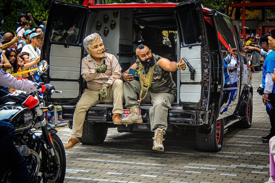 A man with dark hair, wearing sunglasses, a brown leather jacket, and light grey jeans, is sitting on the ground next to a black Royal Enfield motorcycle in a wooded area with tall trees and green foliage. The motorcycle is parked on a patch of grass with its sidestand down, featuring shiny metallic components, a black leather seat, and orange indicator lights. The scene appears to be in natural daylight, with soft shadows cast by the surrounding trees. The man has his hands clasped, looking slightly to the side, and the motorcycle is upright, ready for transport or storage. This setting showcases a casual outdoor environment, possibly related to the preparation or maintenance of a motorbike as part of a house removal or relocation process, as supported by the context of professional removal services offered by Man with Van Enfield.