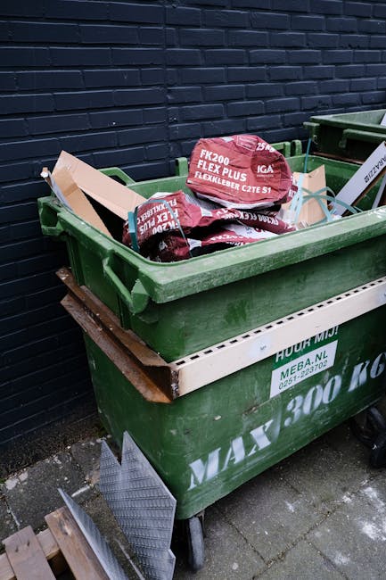 A green wheeled waste bin filled with several red and black packaging bags, some labeled with construction and safety information, and cardboard pieces, positioned against a dark brick wall. The bin i