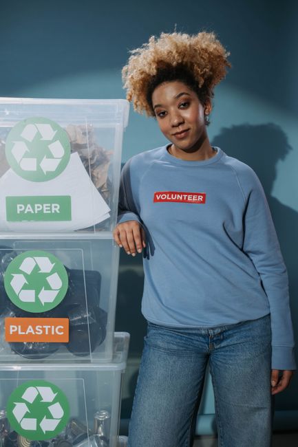 A green wheeled waste bin filled with several red and black packaging bags, some labeled with construction and safety information, and cardboard pieces, positioned against a dark brick wall. The bin i