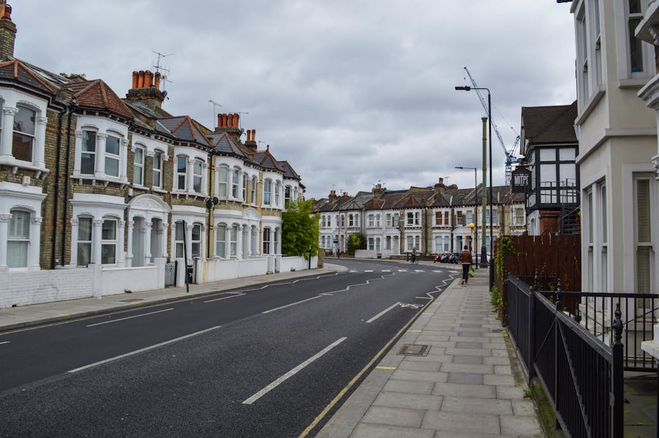 A quiet residential street in Enfield with a row of Victorian-style terraced houses featuring bay windows and brick exteriors, some with white painted facades. The pavement runs along the right side of the image, bordered by black metal railings and a wooden fence, with a few pedestrians visible in the distance. Overhead, street lamps and utility poles line the street, which is partially blocked by a construction crane visible further down. The sky is overcast with grey clouds, indicating typical weather for a house move or relocation process. This scene illustrates the typical environment where Man with Van Enfield provides house removals and local furniture transport services, with the street setting serving as either the starting point or destination for a home relocation or packing and moving operation within the neighbourhood.