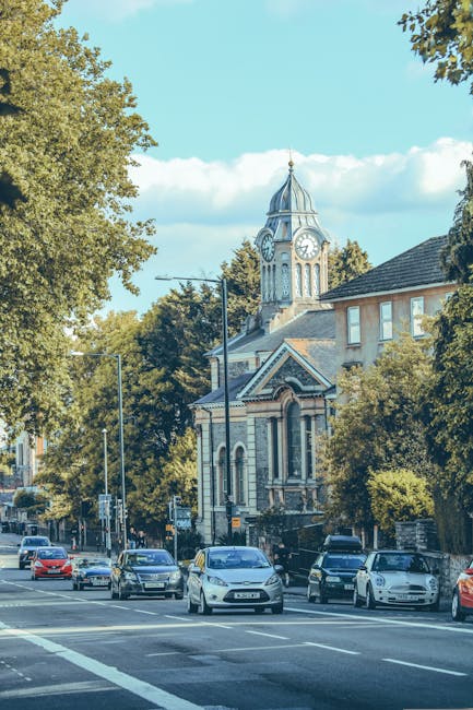 An urban street scene showing a line of parked cars along the pavement in front of a historic building with a prominent clock tower and a decorative dome, surrounded by tall leafy trees under a bright blue sky with scattered clouds. The image captures the move-in or home relocation process, with a focus on the transportation and packing aspects involved in house removals. The setting appears to be a residential or town area in Enfield, with the street likely used for loading or unloading furniture and boxes when relocating. Within the scene, a section of a van or removal vehicle may be visible nearby for loading items, supported by the expertise of Man with Van Enfield. The environment suggests a peaceful and well-established neighbourhood, suitable for house removals, with clear daylight enhancing visibility of the scene.
