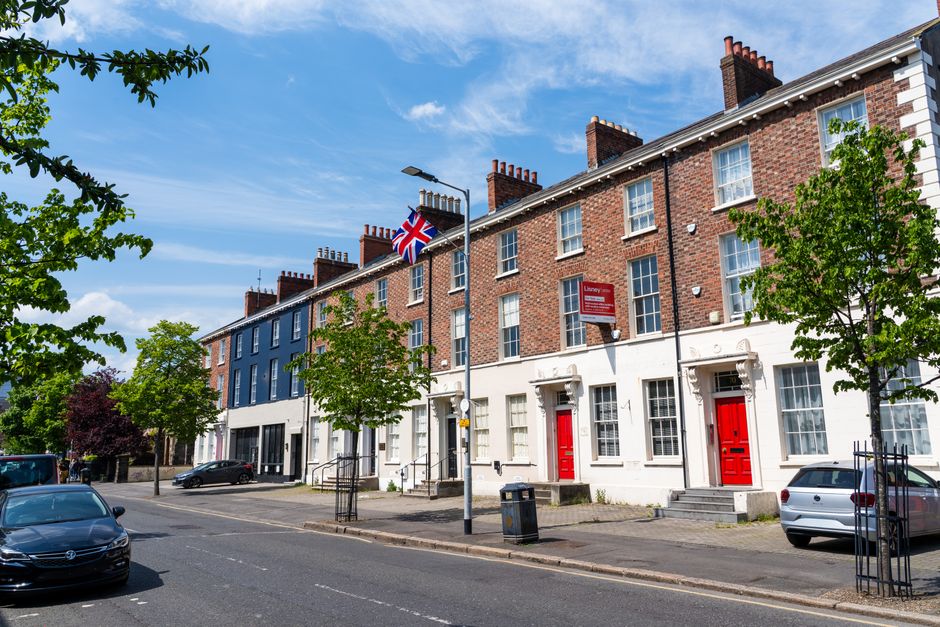 A quiet residential street in Enfield with a row of Victorian-style terraced houses featuring bay windows and brick exteriors, some with white painted facades. The pavement runs along the right side of the image, bordered by black metal railings and a wooden fence, with a few pedestrians visible in the distance. Overhead, street lamps and utility poles line the street, which is partially blocked by a construction crane visible further down. The sky is overcast with grey clouds, indicating typical weather for a house move or relocation process. This scene illustrates the typical environment where Man with Van Enfield provides house removals and local furniture transport services, with the street setting serving as either the starting point or destination for a home relocation or packing and moving operation within the neighbourhood.